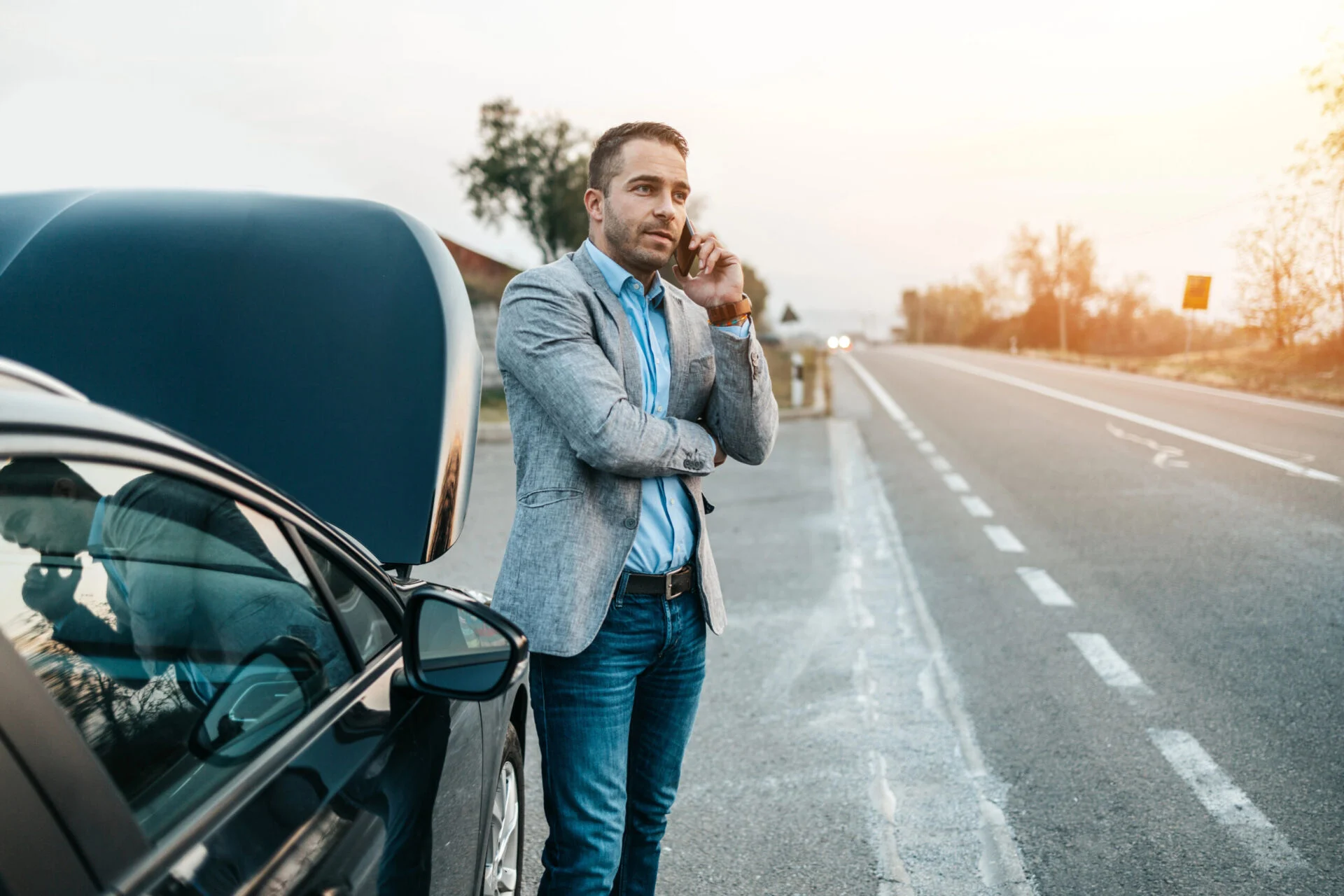 Man talking on a cell phone with the hood of his car popped open on the side of the road