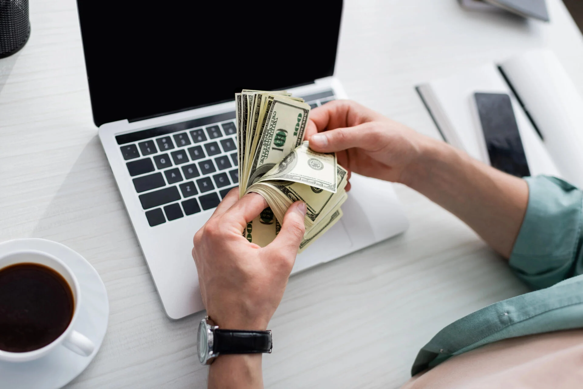 A person thumbs through a stack of money while sitting at a desk with coffee and a laptop