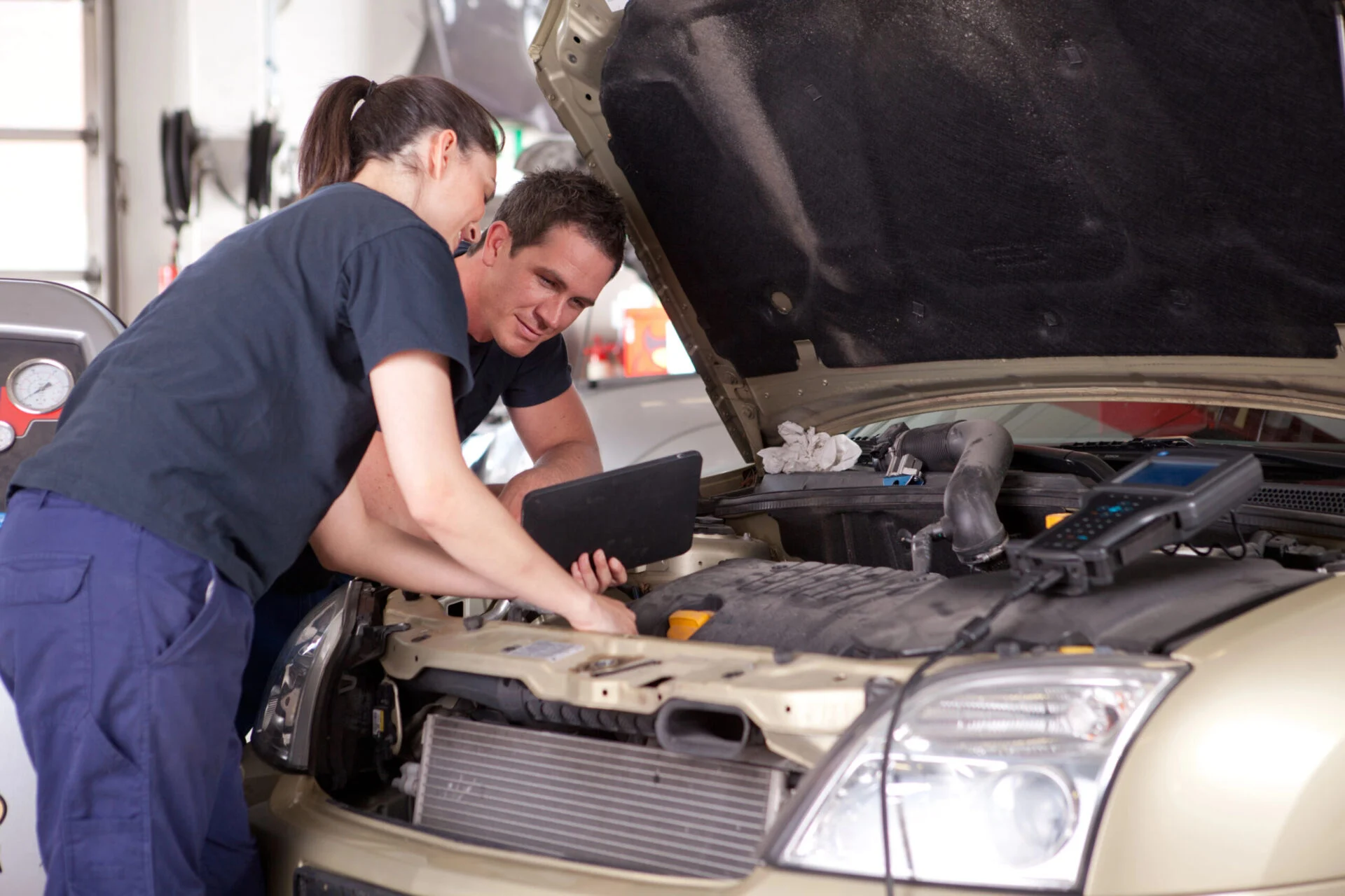 Two mechanics look at a tablet while inspecting under the hood of a vehicle